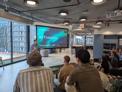 A presenter stands next to a large projection screen displaying 'AI Tinkerers' and 'POLARIS' during an indoor meetup event, with an audience seated in the foreground watching.