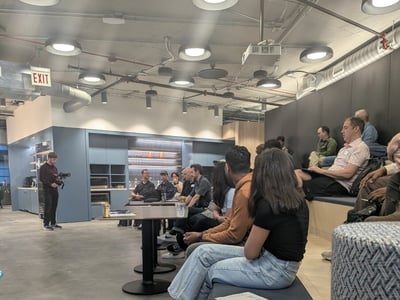 An indoor photograph showing an audience seated on tiered benches and at tables in a modern office or event space, listening to a presentation while a videographer stands to the left.