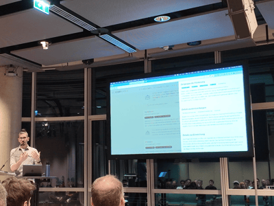 A man is giving a presentation in a modern indoor venue at night, standing at a lectern next to a large screen displaying German text to an audience.
