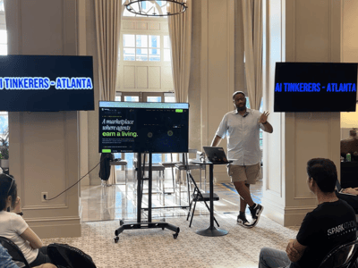 A man is giving a presentation to an audience in a spacious room with high ceilings and large windows, featuring multiple screens displaying event-related text.
