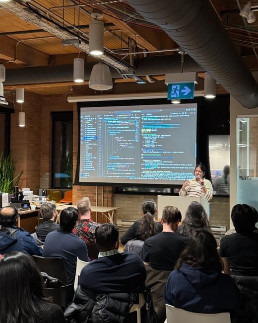 A woman stands at a podium giving a technical presentation to an audience, with a large screen behind her displaying computer code and terminal output.
