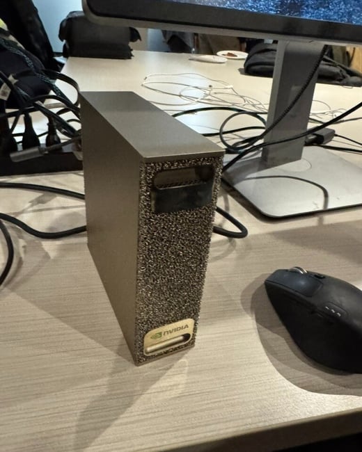 A close-up photograph of a small, textured, bronze-colored computer device, likely an NVIDIA product, resting on a light wooden desk next to a black computer mouse. The background shows a monitor displaying static or a dark pattern, along with various cables and desk clutter.