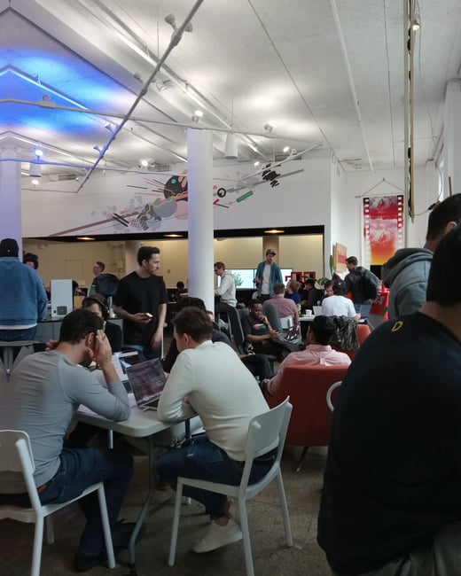 A busy indoor co-working or office space with many people working on laptops at tables and socializing in a modern, high-ceilinged environment.