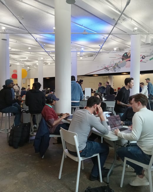 A group of people are working on laptops in a modern, open-plan indoor space with high ceilings and large white pillars.