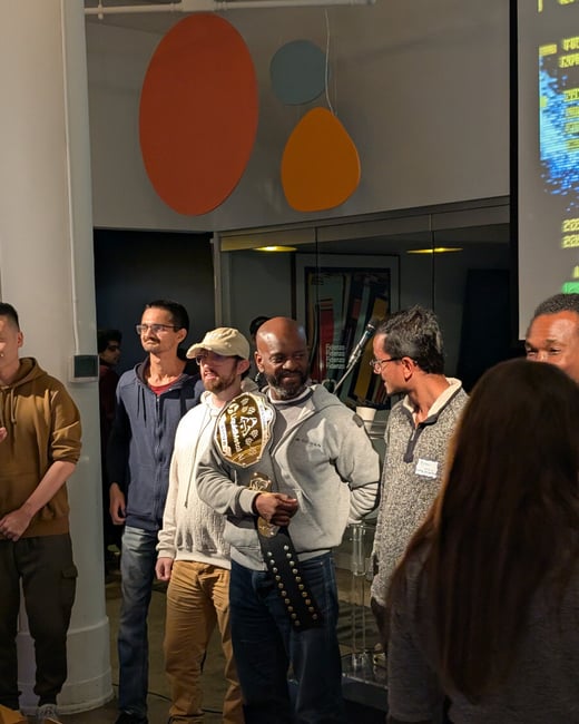 A group of people is gathered indoors at an event, focusing on a man in a grey hoodie who is holding a large championship belt. Abstract colorful shapes decorate the wall behind them, and a projection screen with data is visible on the right.