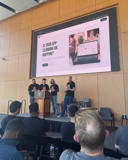 A photograph capturing an indoor business presentation or conference featuring four men on a stage in front of a large screen displaying a website for the product 'APP STATS.' The image is taken from the perspective of the audience, showing the backs of several seated attendees in the foreground.