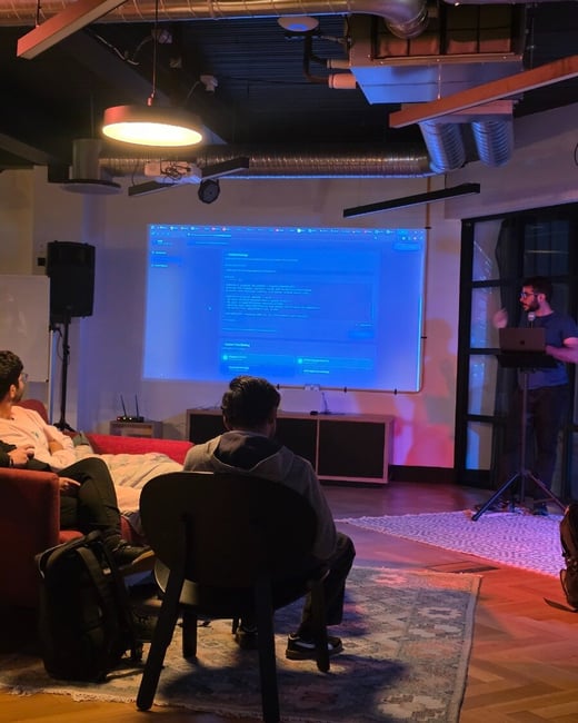 A presenter stands next to a large projection screen displaying a software interface, while audience members watch from chairs and a sofa in a dimly lit, modern event space.