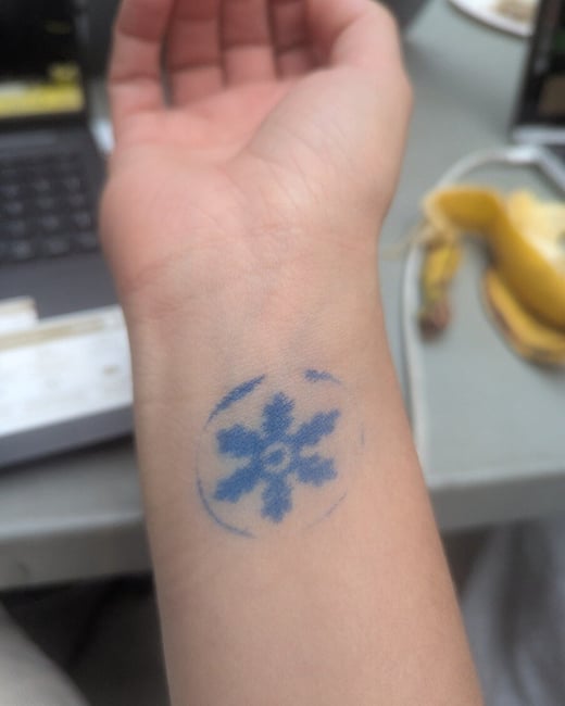 A close-up photograph of a person's forearm and wrist, featuring a temporary blue stamp shaped like a six-pointed snowflake or star inside a faint circle. The background is blurred, showing parts of a laptop keyboard and a peeled banana on a table.