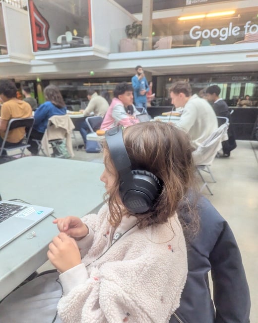 A candid photograph captures a young person wearing black JBL headphones and a fuzzy beige jacket, sitting at a table with an open laptop in a large, busy indoor atrium.