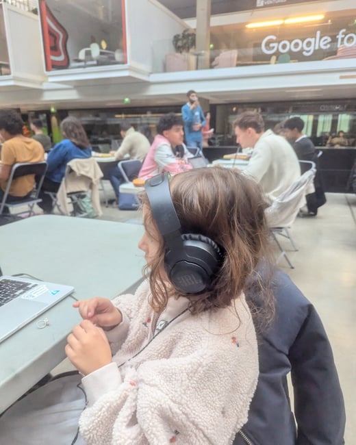 A candid photo shows a young person wearing black JBL headphones and a fluffy light-colored jacket, seated at a table and looking toward a laptop screen in a large, busy indoor space. In the background, many other people are seated at tables, suggesting a co-working session or tech event, with signage for 'Google for VENTURES' visible on an upper level.