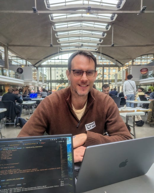 A smiling man wearing glasses and a brown sweater is seated at a table with two laptops, one displaying code, in a large, crowded indoor venue with a glass ceiling.