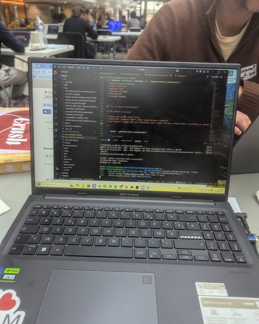 A close-up photograph of a black ASUS Vivobook laptop displaying code and terminal output on a dark background, set within a busy indoor environment like a hackathon or conference workshop.