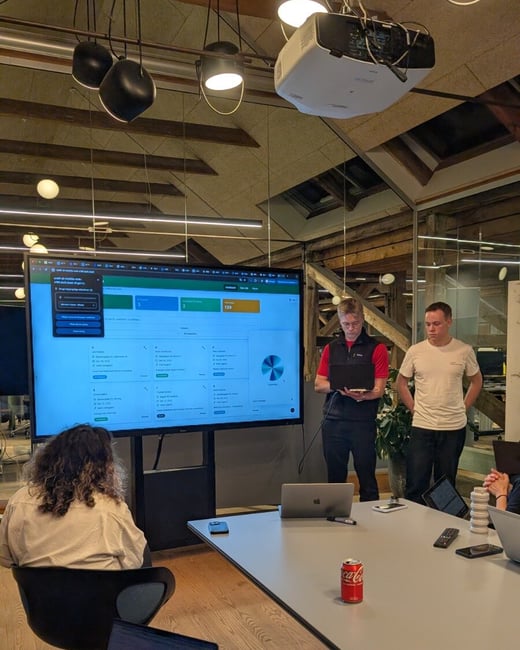 A candid photograph taken during an indoor business meeting or presentation, showing two men standing near a large screen displaying a dashboard while several attendees sit around a table, some working on open laptops.