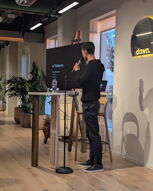 A man wearing a black hoodie and dark pants is giving a presentation indoors, holding a microphone while standing next to a screen displaying the talk title and speaker name.