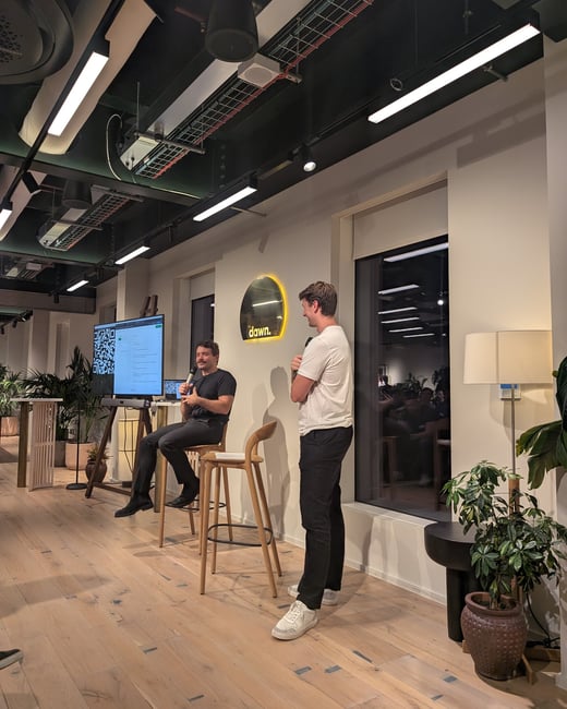 A photograph capturing an indoor panel discussion or presentation featuring two men in a modern, well-lit event space with light wooden floors and an exposed black ceiling. One man is seated on a stool next to a large screen display, while the other stands beside him, near a wall featuring a circular illuminated sign that reads 'dawn.'.