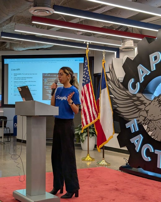 A woman wearing a blue Google shirt stands behind a podium, holding a microphone, and giving a technical presentation in an indoor event space.