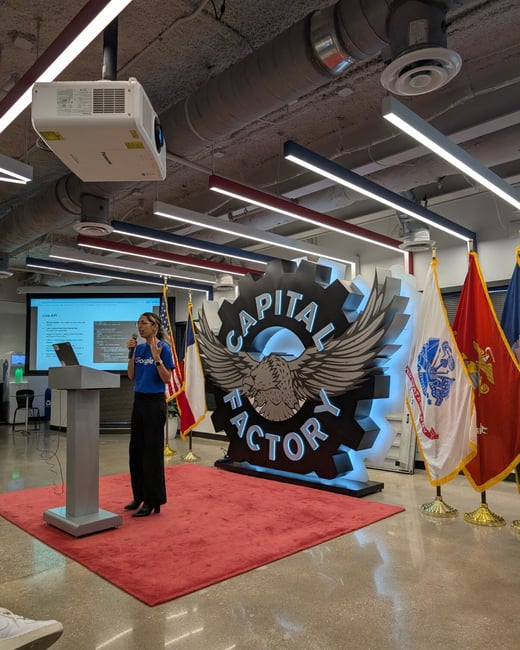 A woman wearing a blue shirt with a Google logo is giving a presentation from a podium on a red rug in a large indoor venue. Behind her is a prominent, illuminated sign featuring an eagle and the text "CAPITAL FACTORY," alongside several flags.