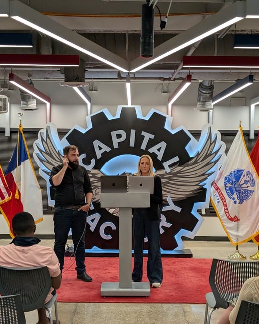 A photograph captures an indoor presentation where a man and a woman stand behind a podium with laptops, addressing an unseen audience. The backdrop features a large, stylized gear logo for 'Capital Factory' flanked by the French and US Army flags.