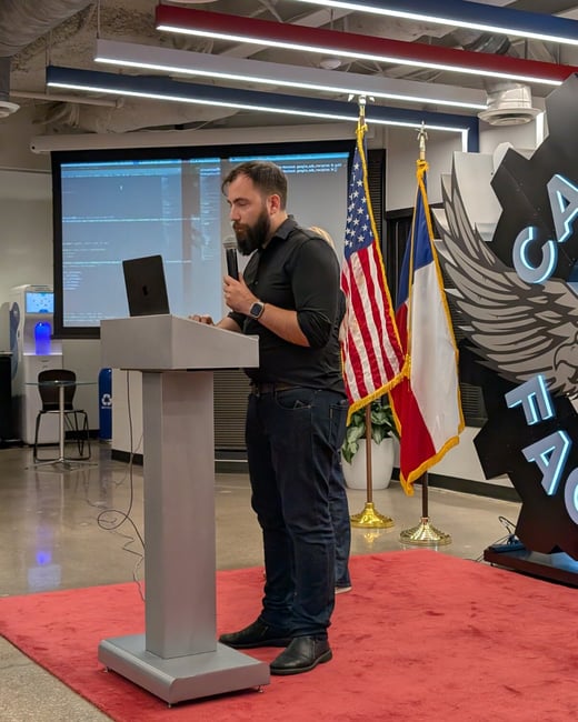 A bearded man in a black shirt stands at a silver podium, holding a microphone and interacting with a laptop, while giving a technical presentation indoors.