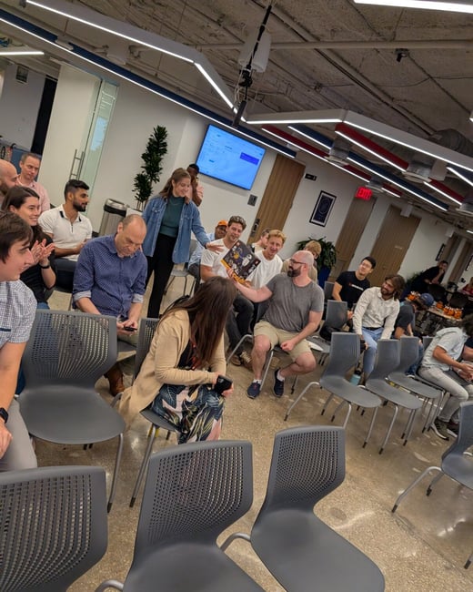 A candid photograph captures a large group of people gathered in a modern office space, sitting in rows of chairs during what appears to be a presentation or group discussion.