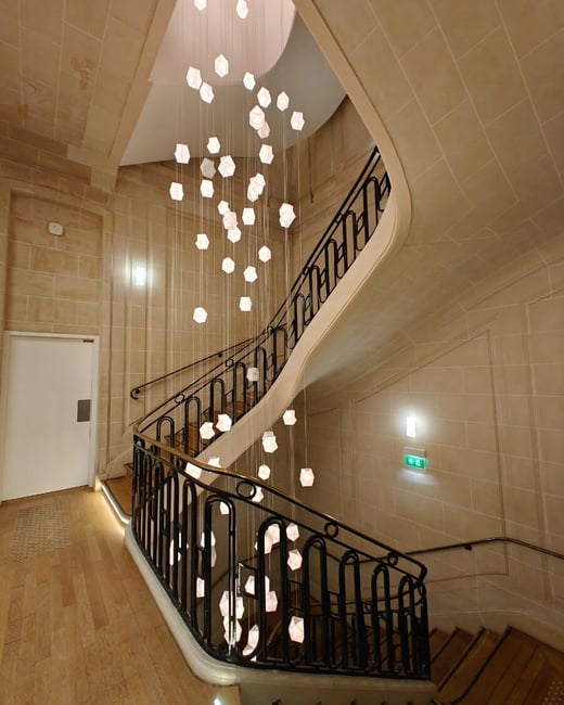 A photograph showing an elegant, curving staircase within a classical building, featuring light tan stone walls and a dark iron railing. A dramatic, multi-story chandelier composed of numerous suspended geometric light fixtures hangs in the center of the stairwell.