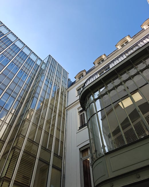 A low-angle photograph capturing the striking architectural contrast between a modern glass skyscraper and a traditional white building featuring a curved glass and metal bay window, all set against a clear, bright blue sky.