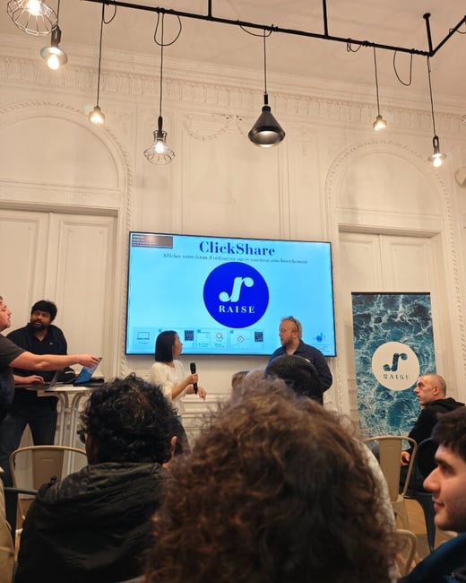 A photograph capturing an indoor presentation or meeting, showing a woman speaking into a microphone in front of a large screen that displays the 'ClickShare' and 'RAISE' logos, while an audience watches.