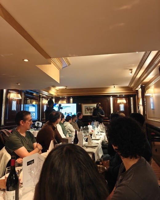 A group of people is gathered around a long dining table in a wood-paneled room, attending a presentation or dinner meeting. A speaker stands at the far end of the table, with a large screen behind him displaying the text "MULTIMODAL AI."