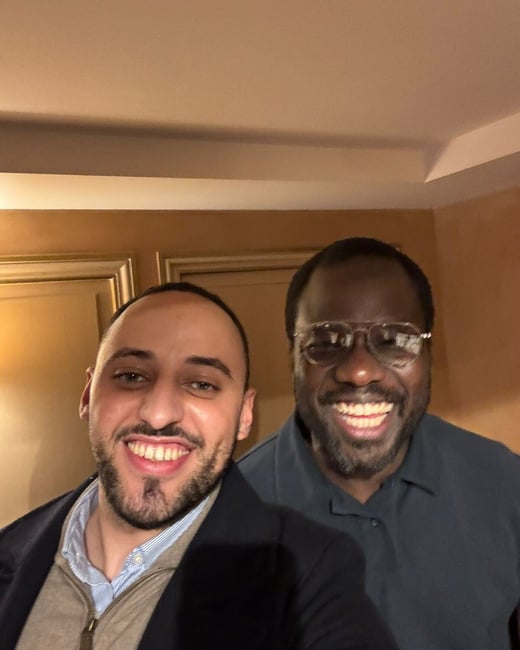 Two men are taking a close-up selfie indoors, both smiling broadly and looking directly at the camera.