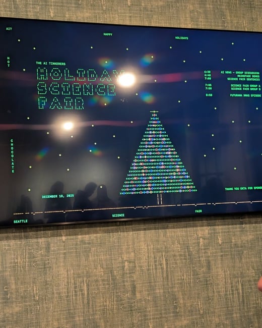 A photograph of a large monitor displaying a retro, green ASCII art-style graphic advertising 'The AI Tinkerers Holiday Science Fair' and listing an event agenda. The screen reflects bright overhead lights and the dark silhouettes of several people standing nearby.