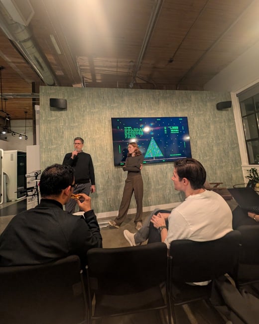 A dimly lit indoor photograph capturing a presentation or event, where two people stand in front of a screen displaying 'HOLIDAY SCIENCE FAIR' while several audience members watch from the foreground.