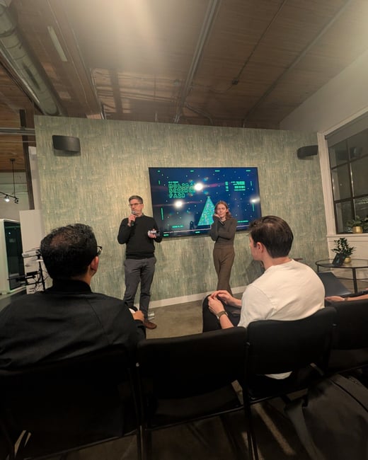 A photograph taken from the audience shows two presenters, a man and a woman, standing in front of a large screen displaying the title "HOLIDAY SCIENCE FAIR" in a dimly lit indoor event space.