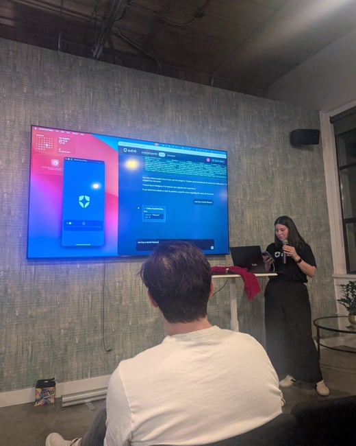 A low-light photograph capturing an indoor technology presentation, showing a woman presenting next to a large screen displaying a mobile and desktop application interface, while an audience member sits in the foreground facing away.