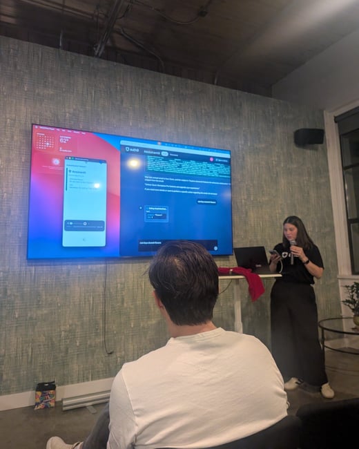 A candid indoor photograph capturing a woman giving a presentation or software demonstration using a large screen, while an audience member is visible in the foreground viewed from the back.