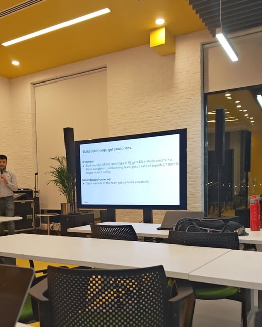 A photograph taken from the audience perspective during an indoor event or presentation, showing a speaker standing next to a large screen displaying prize details for a competition, set against a backdrop of white brick walls and a bright yellow ceiling.