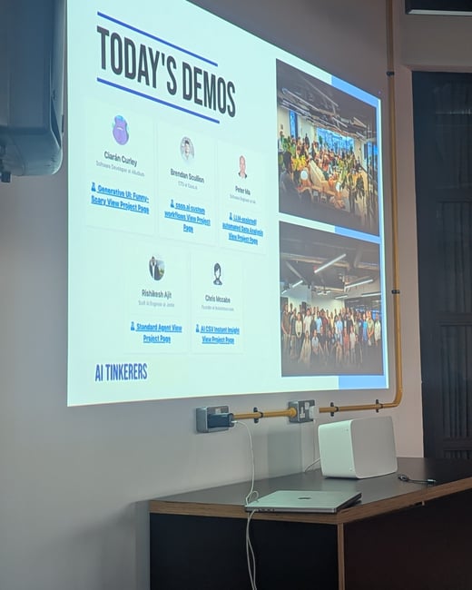 A photograph of a projected presentation slide titled "TODAY'S DEMOS," listing five presenters and their AI-related projects, taken in an indoor setting with a laptop resting on a dark desk below the screen.