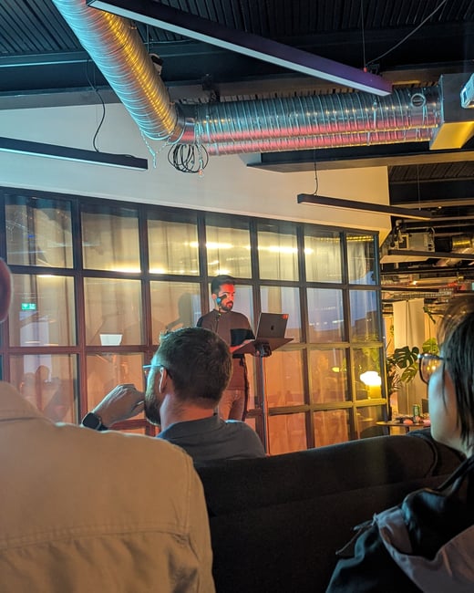 A low-light photograph of an indoor event where a speaker, standing with a laptop, is giving a presentation to a seated audience in a modern venue with exposed industrial ductwork and large glass window panels.