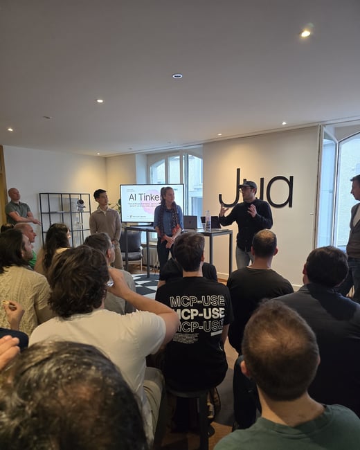 A group of people are gathered in an indoor space for a presentation titled 'AI Tinkerers'. A man in a cap is speaking to the audience while standing near a screen and a large 'Jua' logo on the wall.