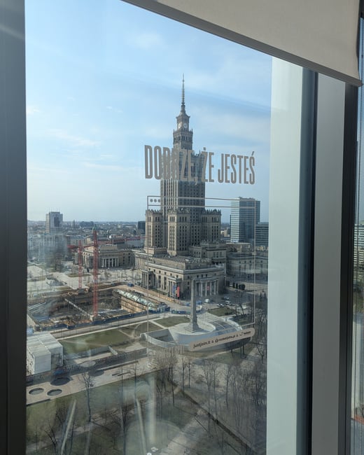 A view of the Palace of Culture and Science in Warsaw, Poland, seen through a window with a decal that reads 'DOBRZE, ŻE JESTEŚ'.