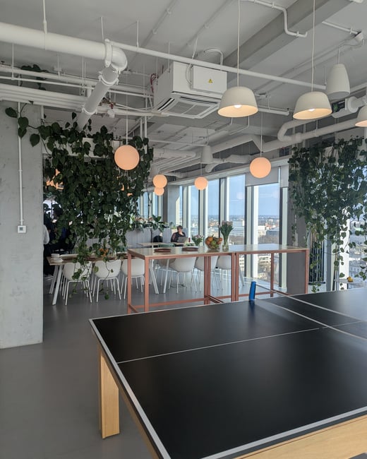 A modern, bright office common area featuring a ping pong table in the foreground, dining tables, and lush hanging plants with a city skyline view through large windows.