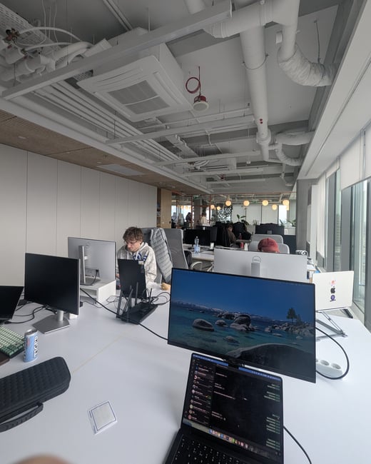 A wide-angle shot of a modern open-plan office with employees working at desks featuring multiple monitors and laptops under an industrial-style exposed ceiling.
