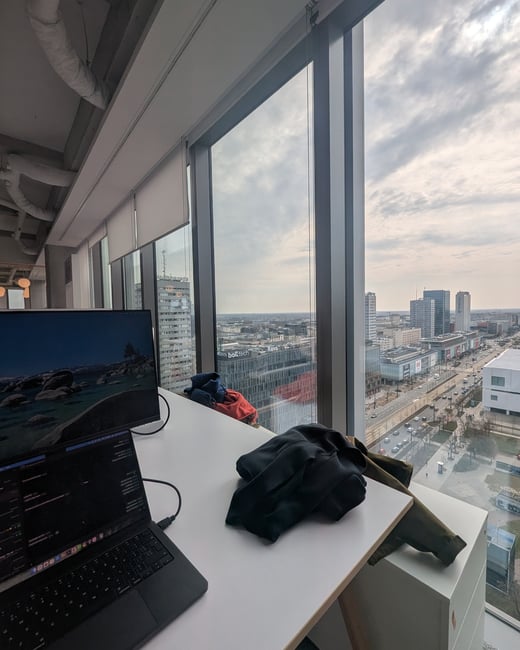 A view from a high-rise office window showing a white desk with a laptop and monitor, overlooking a cloudy cityscape.