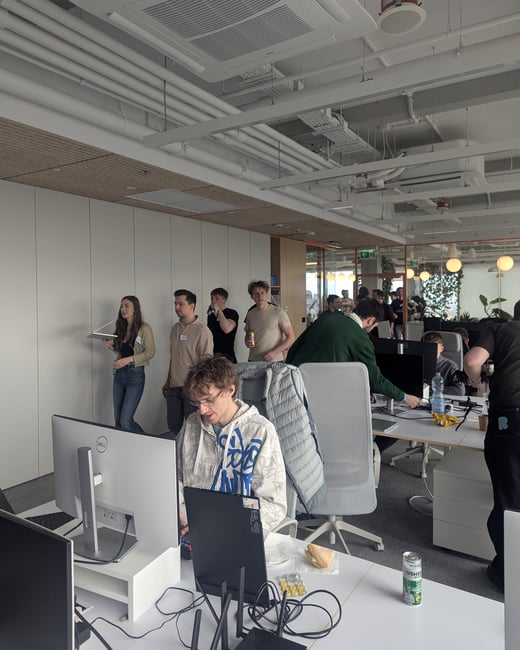 A group of young people are gathered in a modern, open-plan office space with exposed ceiling pipes, some standing and talking while others work at desks with computers.