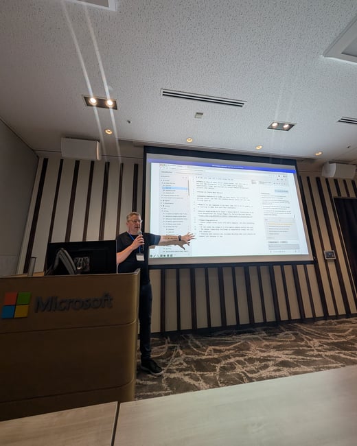 A presenter stands on a stage next to a Microsoft-branded podium, gesturing towards a large projector screen displaying text and code.
