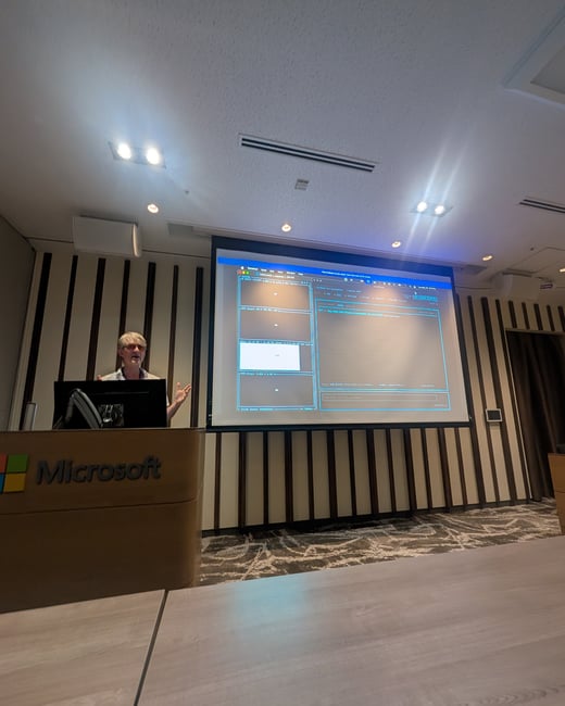 A presenter stands at a Microsoft-branded wooden podium, gesturing towards a large projection screen displaying a terminal interface.