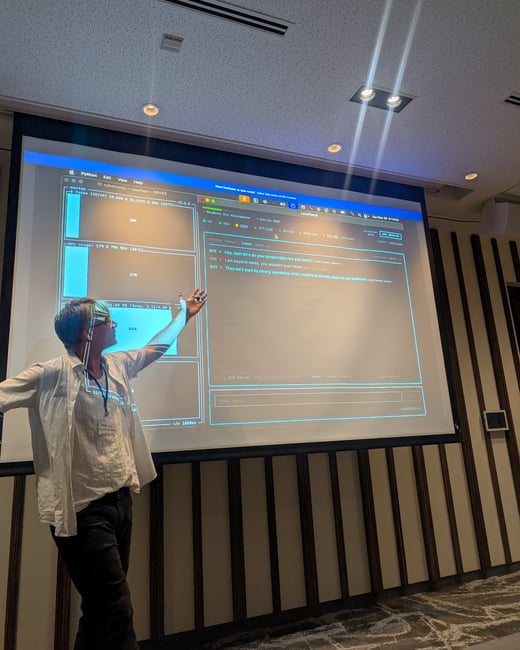 A person stands in front of a projection screen displaying a terminal interface, gesturing towards the screen during a presentation.