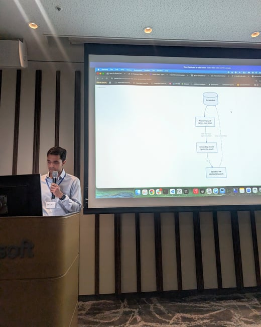 A man stands at a podium holding a microphone while presenting next to a large projector screen displaying a technical flowchart.
