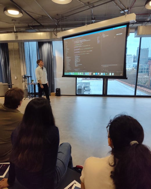 A man stands in a modern office space giving a coding presentation on a large projection screen to an audience sitting on the floor.