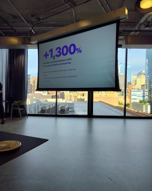A photograph of a presentation screen inside a modern room with large windows overlooking a city skyline. The screen displays a slide showing a statistic of '+1,300%' regarding deepfake voice fraud attempts in 2024.