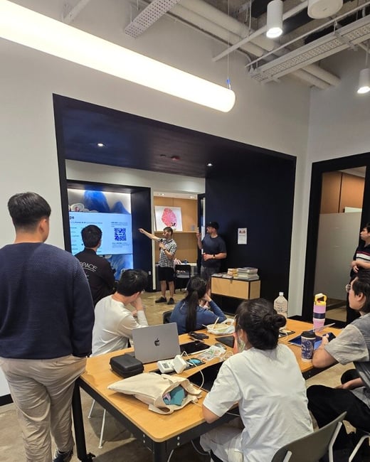 A candid indoor photo capturing a group of people attending a presentation or meeting in a modern office space, where a speaker points toward a large screen displaying information and a QR code.
